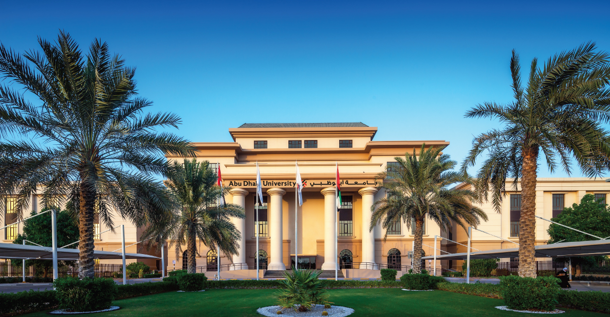 Abu Dhabi University main building with flags and palm trees under clear blue sky.