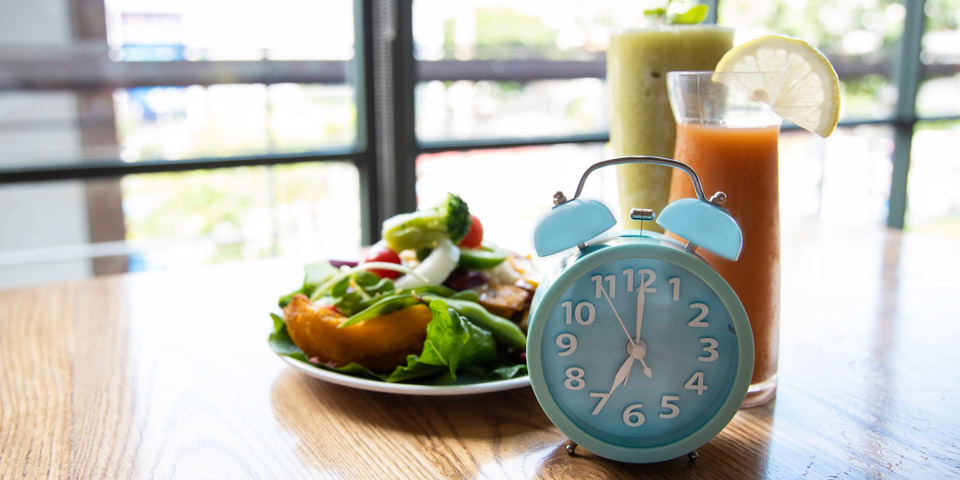 Clock in front of a plate of food and glass of juice representing intermittent fasting